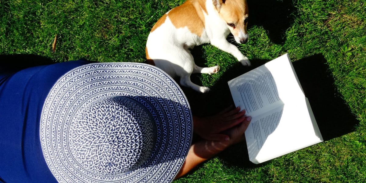 lady in large hat laying on her front on the lawn reading a book with her dog beside her