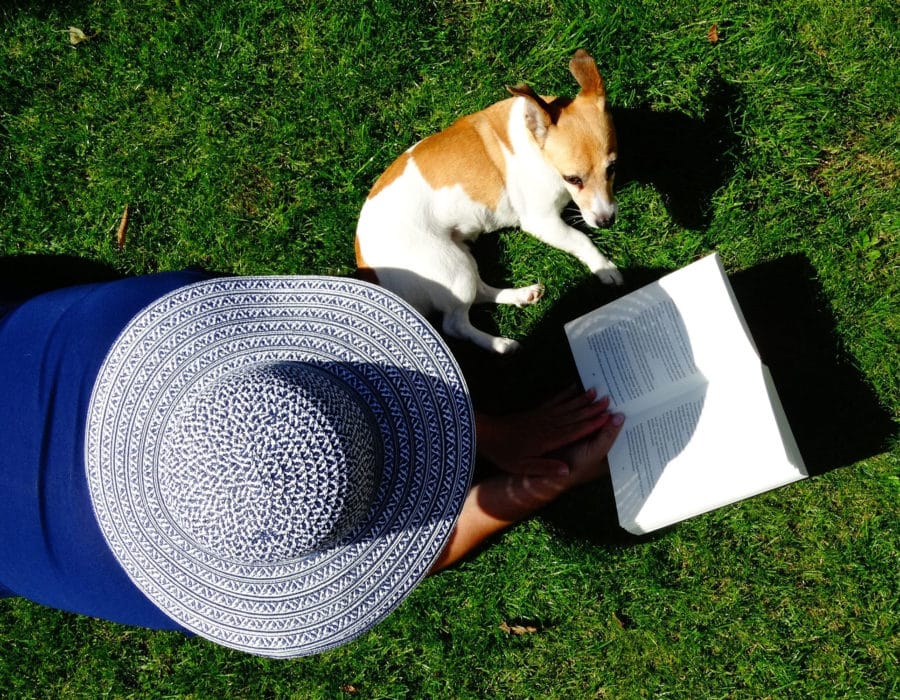 lady in large hat laying on her front on the lawn reading a book with her dog beside her